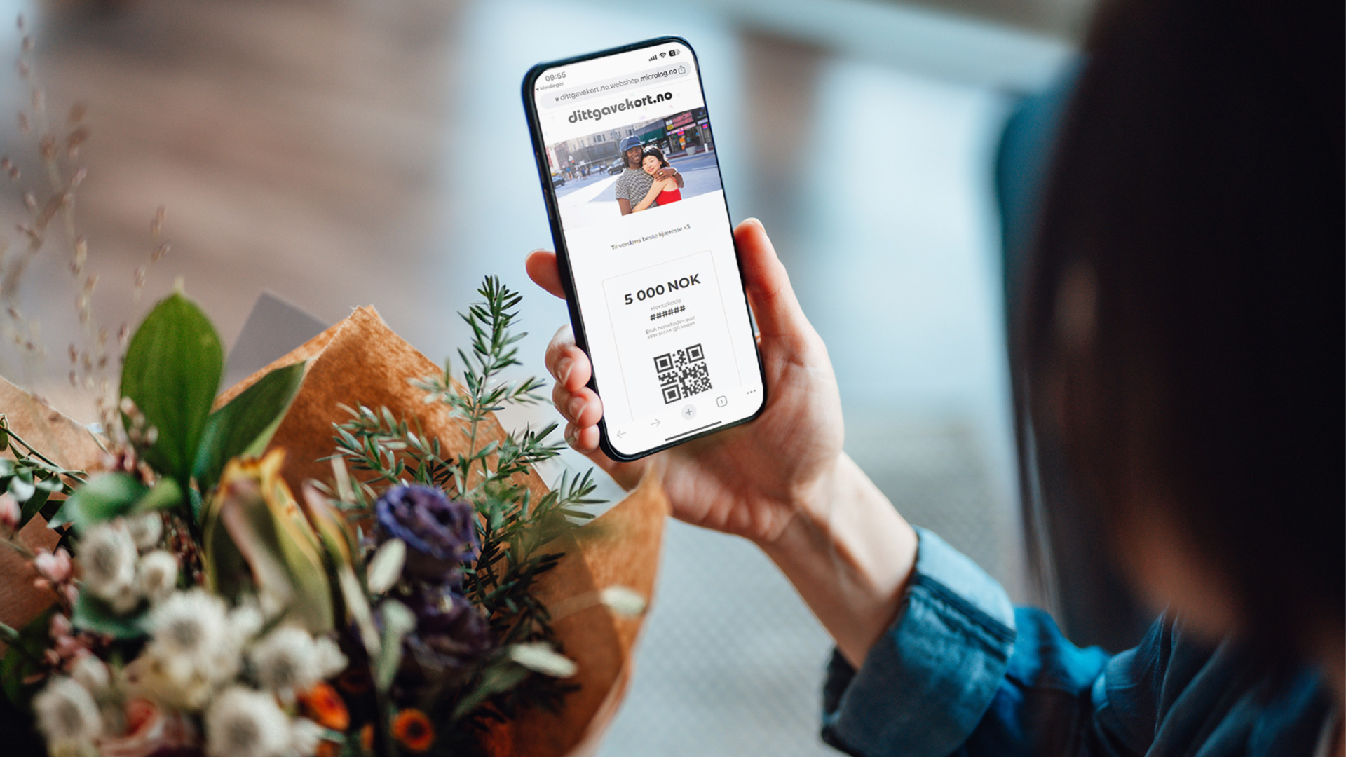 En dame holder mobil med dittgavekort og et bukett med blomster foran seg