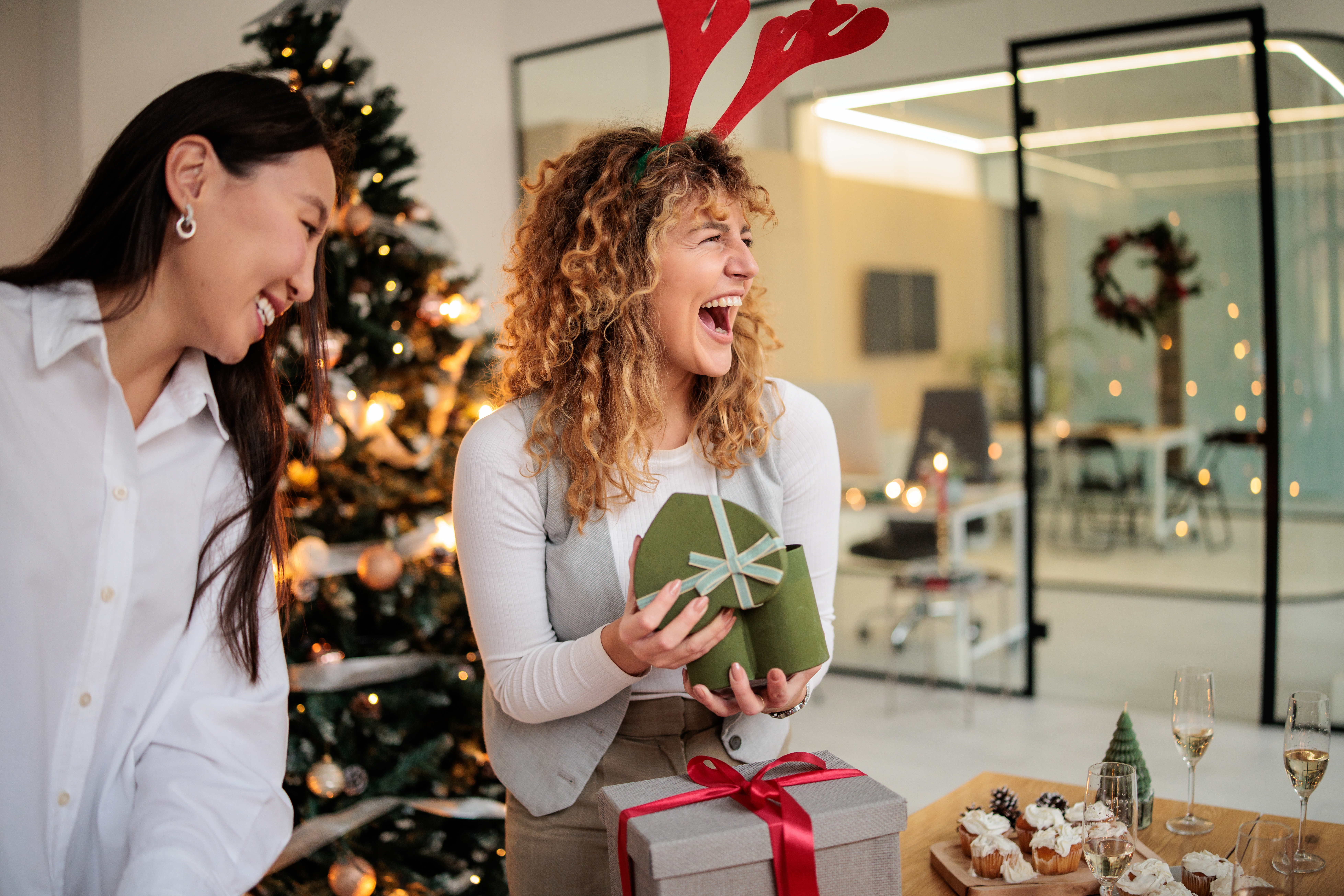 En kvinne åpner en gave mens en annen kvinne står og ser på, med et juletre i bakgrunnen.
