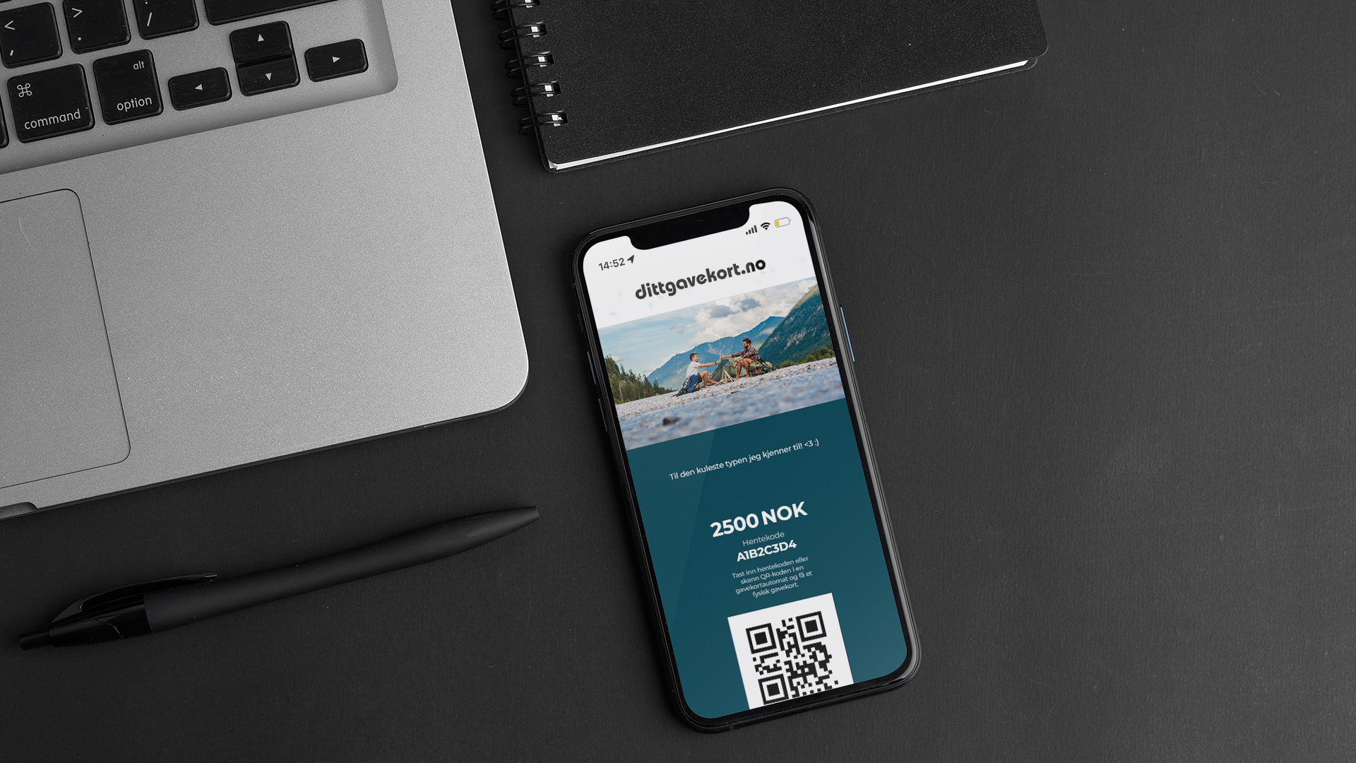 En mobil med dittgavekort ligger ved siden av en PC på et sort skrivebord, sammen med en sort penn og en sort notatbok.